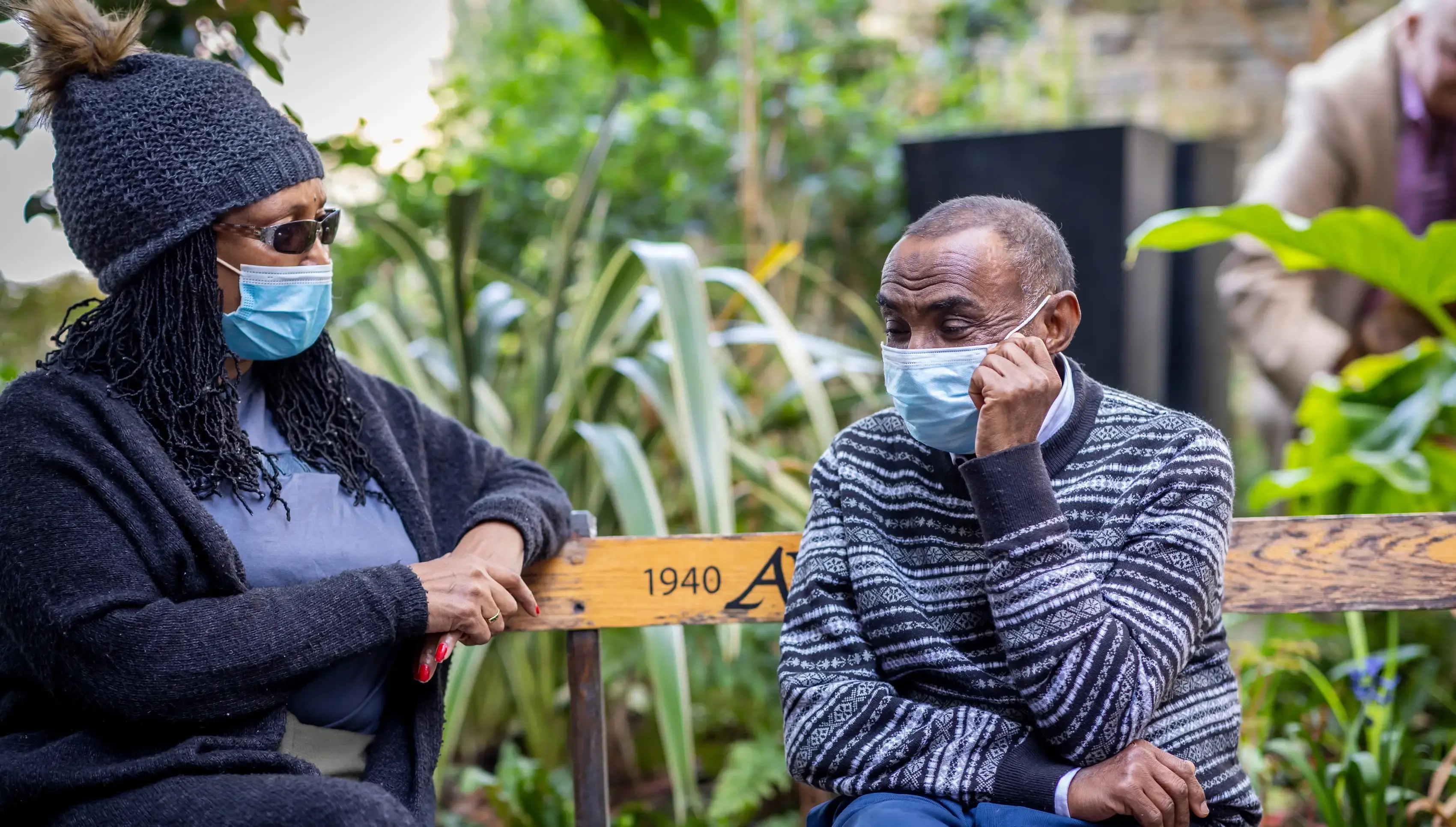 An older adult man and his caregiver sitting in a garden on a wood bench, wearing sweaters and masks.